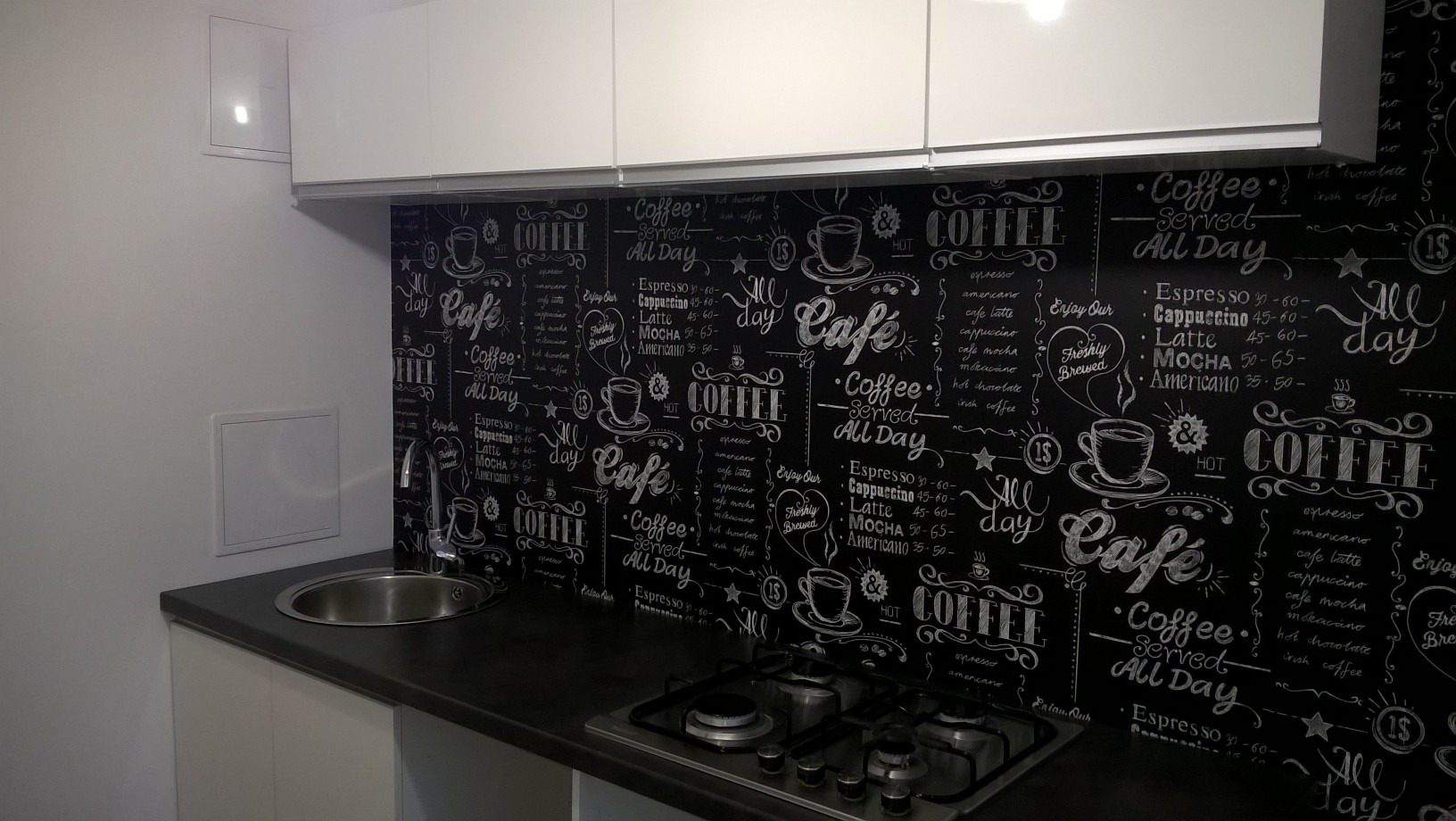 Nowoczesna kuchnia z białymi szafkami i czarnym blatem, nad którym znajduje się dekoracyjna ściana z motywem kawy napisanym kredą.