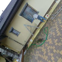 Zaklad-elektrobudowlany - Pracownik maluje elewację domu, okna zabezpieczone folią, widoczna drabina, wiadro z farbą i wąż ogrodowy.