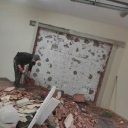 Zaklad-elektrobudowlany - Pracownik w czarnym ubraniu usuwa resztki starej ściany z cegieł, widoczny bałagan po remoncie, gruz na podłodze, puste wiadra po farbie i fragmenty cegieł