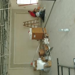 Zaklad-elektrobudowlany - Prace wykończeniowe w nowym mieszkaniu: szkielet ścian działowych z instalacją elektryczną, pracownik szpachlujący ścianę pod oknem, drabina, kartony i materiały budowlane na podłodze, butelka wody...