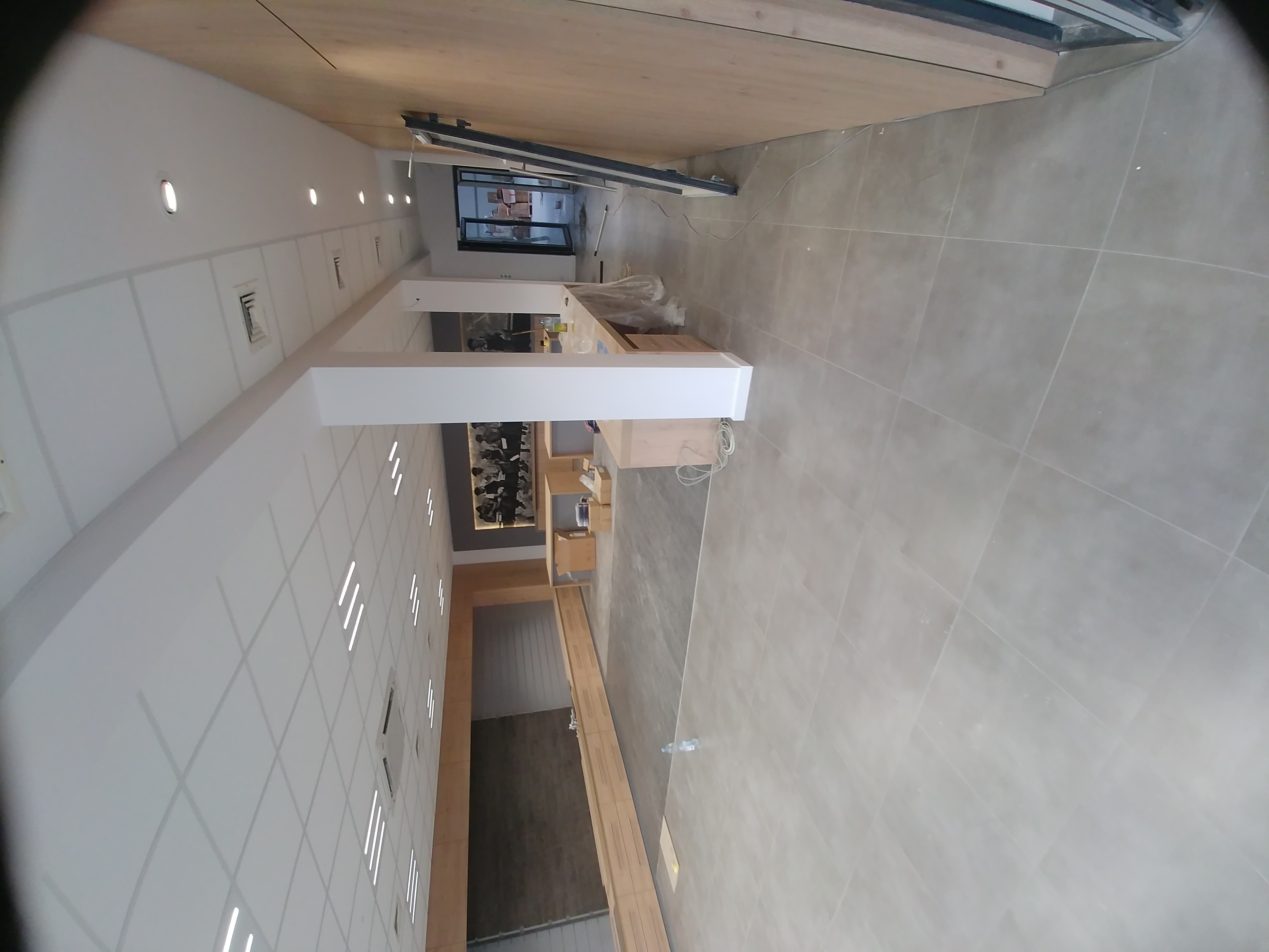 Wnętrze lokalu usługowego w trakcie wykańczania: widoczne płytki podłogowe, ściany z podwieszanym sufitem z oświetleniem punktowym, drewniane wykończenia i częściowo zasłonięte okno wystawowe...