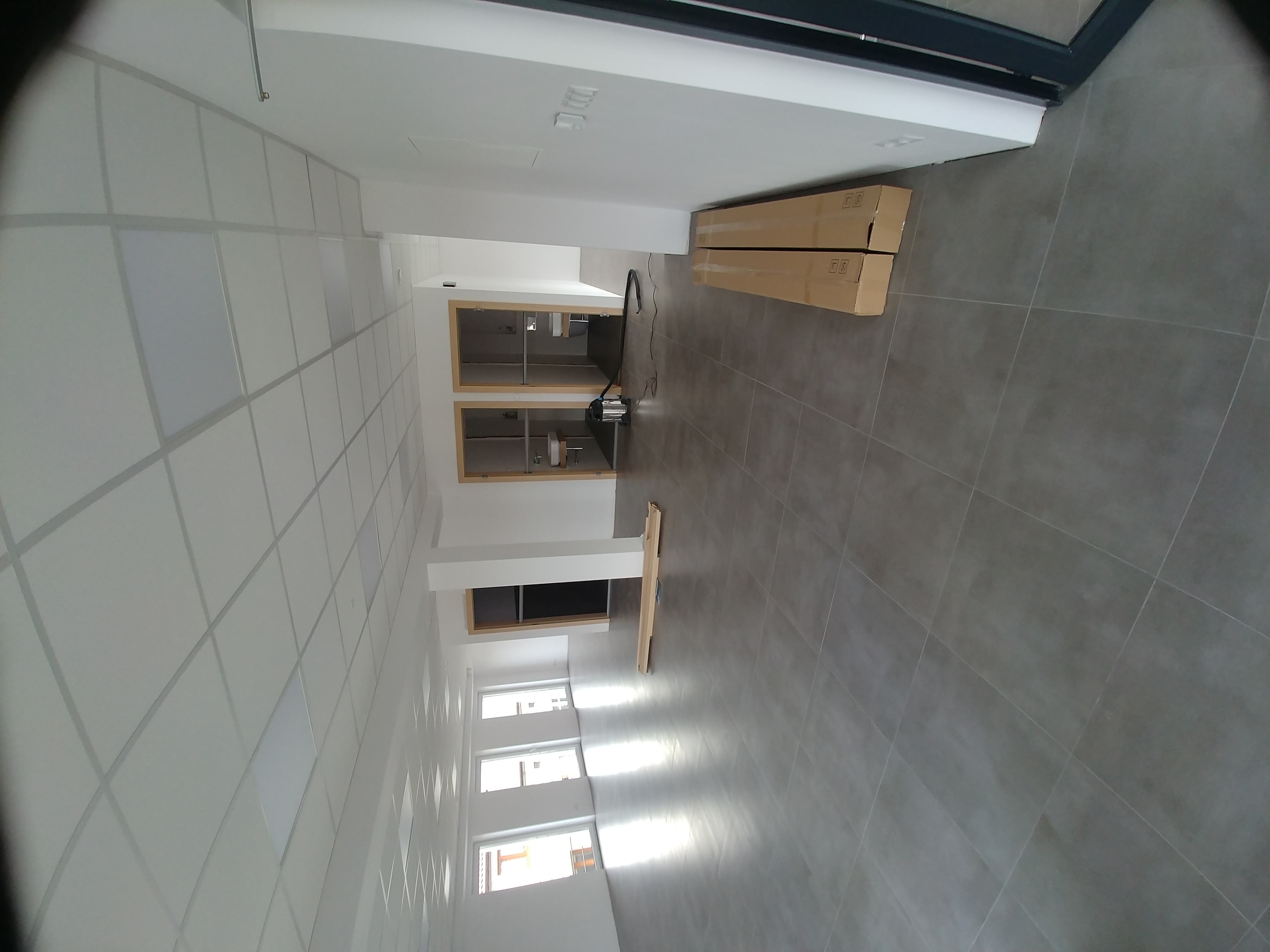 Wnętrze w trakcie wykańczania: szara płytka podłogowa, biały sufit kasetonowy, białe ściany, okna z widokiem na zewnątrz, kartony z materiałami budowlanymi oraz odkurzacz przemysłowy.