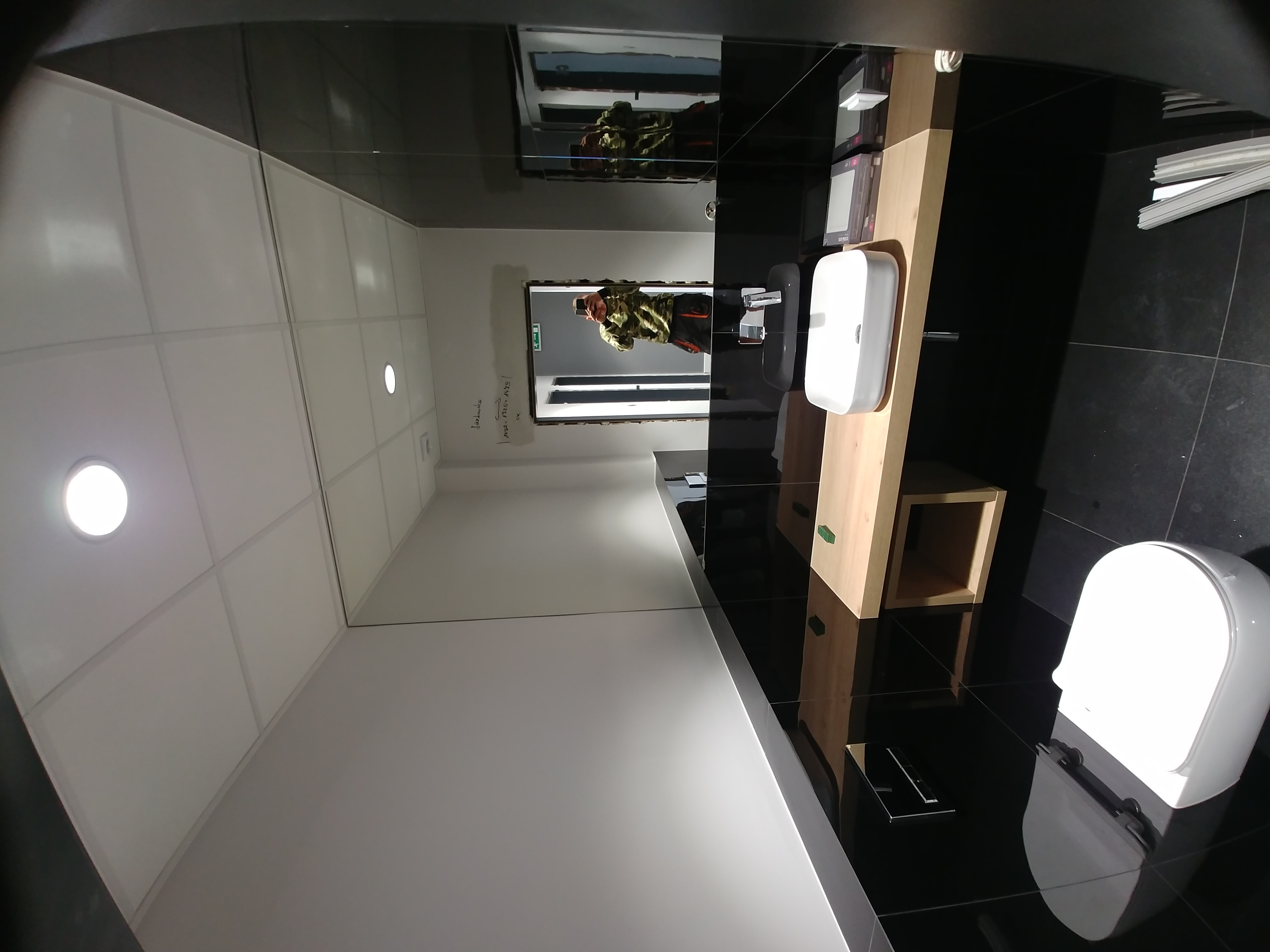 Nowoczesna łazienka z czarnymi płytkami podłogowymi, białą armaturą (toaleta i umywalka na drewnianym blacie), oświetleniem sufitowym i odbiciem osoby robiącej zdjęcie w lustrze.