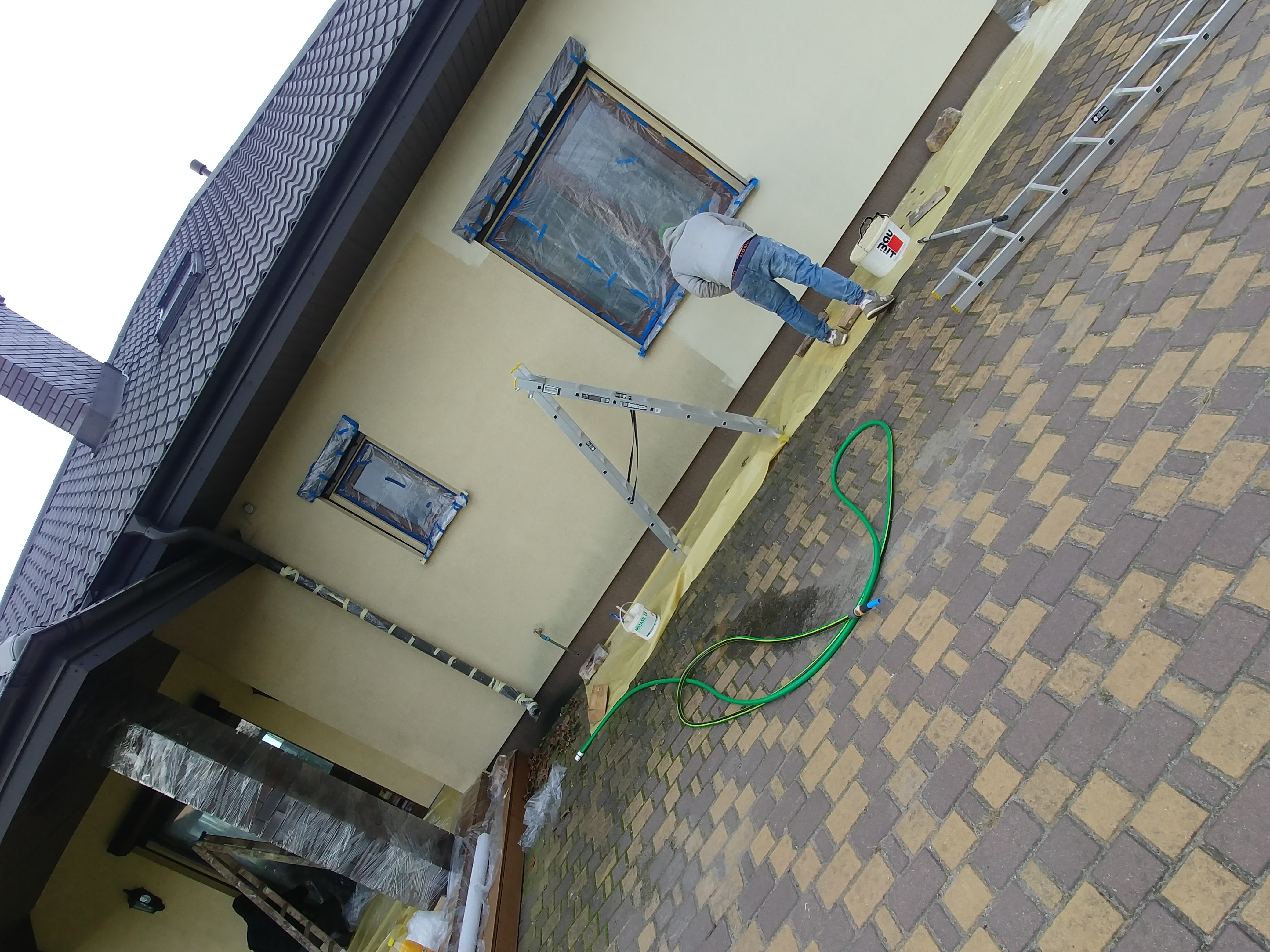 Pracownik maluje elewację domu, okna zabezpieczone folią, widoczna drabina, wiadro z farbą i wąż ogrodowy.