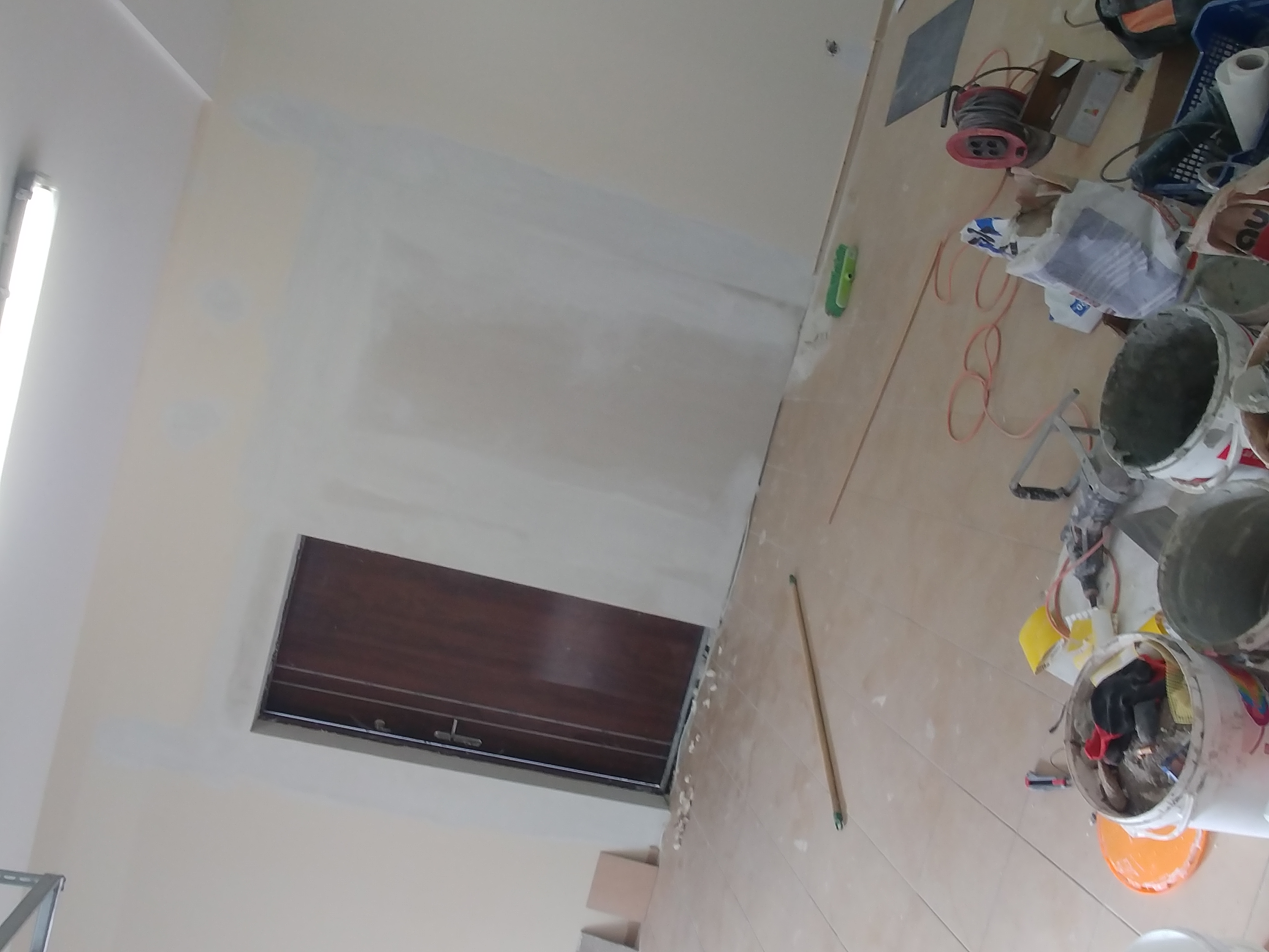 Ściana w trakcie prac wykończeniowych, widoczne ślady po gładzi, narzędzia malarskie i wiadro z farbą na podłodze, brązowe drzwi z metalowym zamkiem w tle.