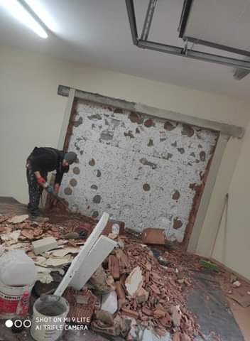 Pracownik w czarnym ubraniu usuwa resztki starej ściany z cegieł, widoczny bałagan po remoncie, gruz na podłodze, puste wiadra po farbie i fragmenty cegieł