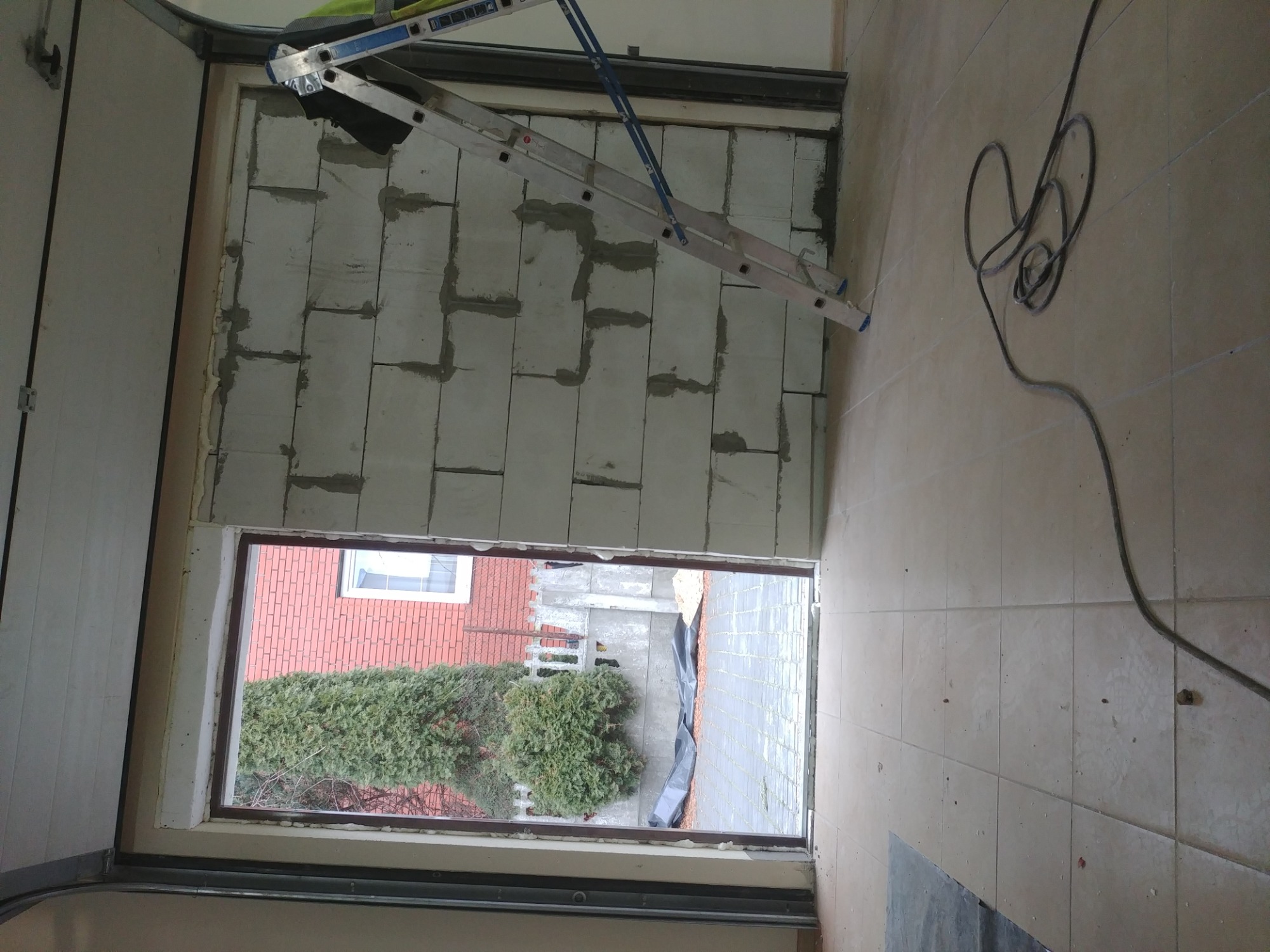 Ocieplanie ściany garażu z widoczną drabiną i kablem elektrycznym na podłodze z płytek, widok przez okno na zewnątrz.