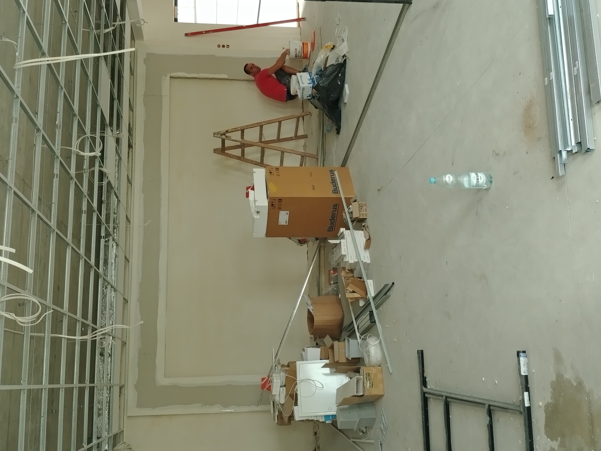 Prace wykończeniowe w nowym mieszkaniu: szkielet ścian działowych z instalacją elektryczną, pracownik szpachlujący ścianę pod oknem, drabina, kartony i materiały budowlane na podłodze, butelka wody...