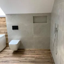 Nowoczesna łazienka z szarymi płytkami imitującymi kamień i drewnopodobną podłogą, wisząca toaleta z czarnym panelem sterującym, deszczownica i odpływ liniowy.