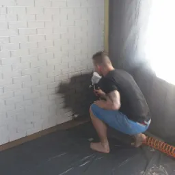 Mężczyzna maluje białą ścianę z cegieł natryskowo na szaro, zabezpieczona folią podłoga i ściany, widoczny kompresor.