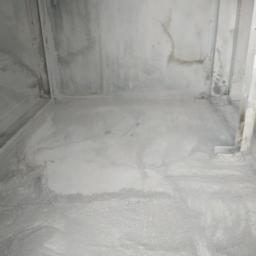 Hydraulik/Monter klimatyzacji - Surowy stan posadzki i ścian w pomieszczeniu przed pracami wykończeniowymi, widoczne ślady wilgoci na ścianach, na podłodze warstwa pyłu i gruzu.