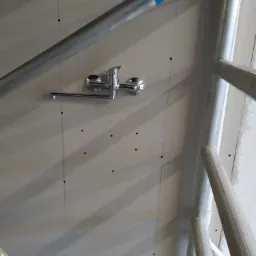 Chromowana bateria kuchenna zamontowana na ścianie, widoczna na tle surowej ściany z widocznymi liniami pionowymi i otworami, otoczona elementami rusztowania.