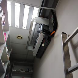 Hydraulik/Monter klimatyzacji - Wnętrze biura z widoczną jednostką wewnętrzną klimatyzacji w trakcie montażu, z otwartą obudową, widoczne okablowanie i drabina przystawiona do ściany. W tle regały z lekami.