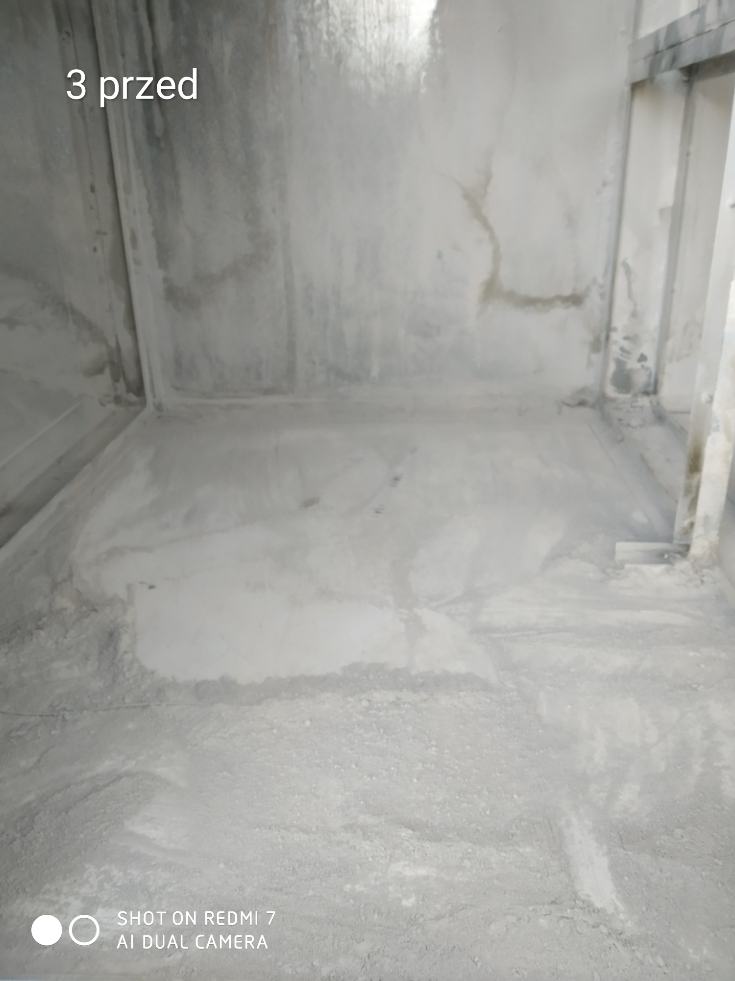 Surowy stan posadzki i ścian w pomieszczeniu przed pracami wykończeniowymi, widoczne ślady wilgoci na ścianach, na podłodze warstwa pyłu i gruzu.