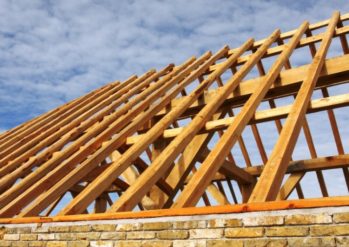 Konstrukcja drewniana dachu w trakcie budowy, widok na krokwie i murłatę z cegły, na tle błękitnego nieba.