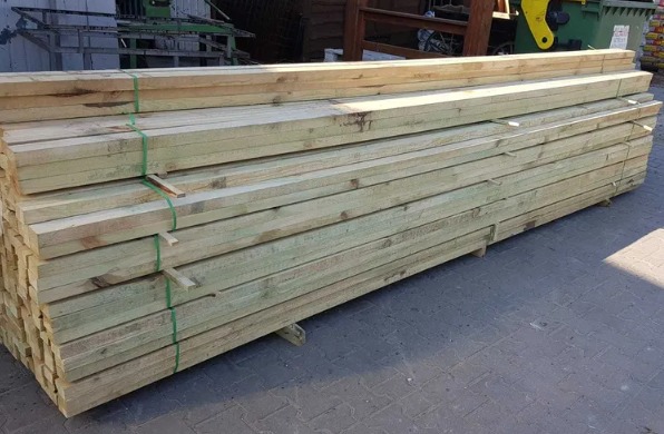 Stos impregnowanych desek budowlanych, zabezpieczonych zieloną taśmą, ułożonych na palecie, widocznych na tle fragmentu placu składowego.
