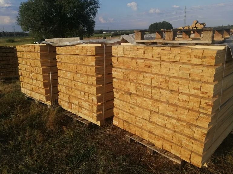 Stosy świeżo przetartej tarcicy sosnowej na paletach, zabezpieczone taśmą, składowane na zewnątrz na trawiastym podłożu, z widocznymi w tle stosami okrąglaków.