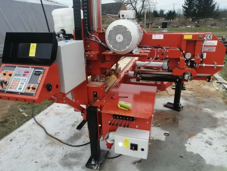 Pomarańczowa, stacjonarna piła taśmowa do drewna w trakcie cięcia deski na zewnątrz, z widocznym panelem sterowania i silnikiem elektrycznym.