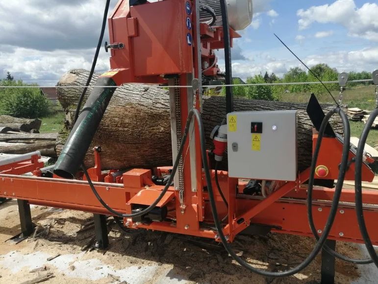 Pomarańczowa trakotarka taśmowa tnie kłodę drewna na deski, widoczne trociny i przewody, na tle zieleni i pochmurnego nieba.