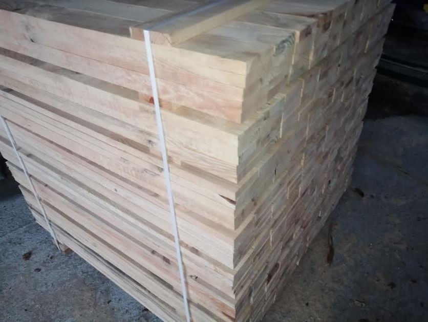 Stos jasnej, świeżo przetartej tarcicy budowlanej, związany białą taśmą, widoczny z boku w magazynie.