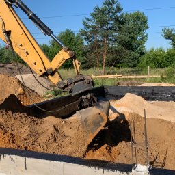 MARMAX - Żółta koparka CAT zasypuje fundament piaskiem. Widoczne zbrojenia i izolacja fundamentu. Prace ziemne w toku, błękitne niebo w tle.