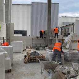 MARMAX - Murowanie ścian z betonu komórkowego na budowie hali. Widoczni pracownicy w odblaskowych kamizelkach i kaskach, rusztowania, palety z bloczkami i narzędzia murarskie.