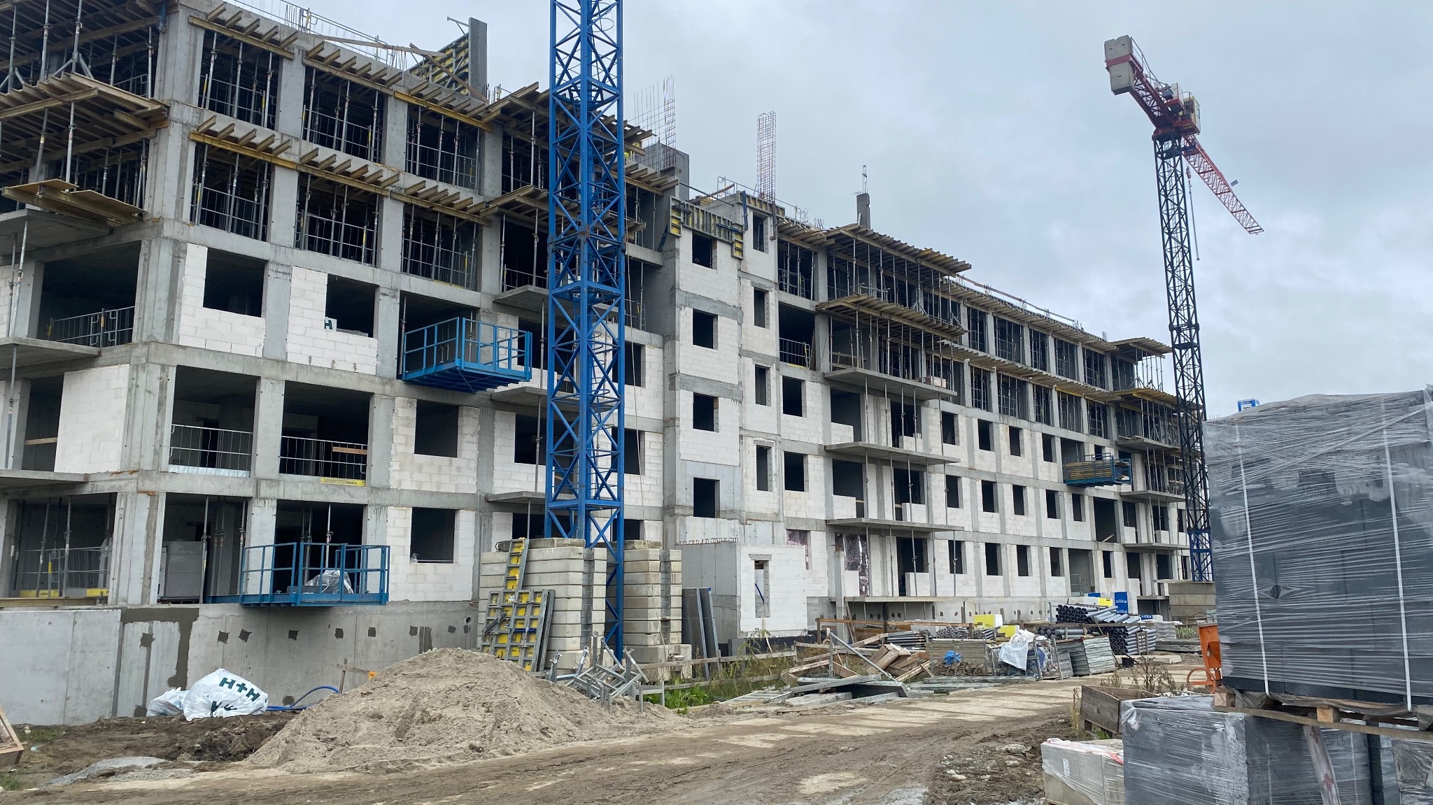 Wznoszenie wielopiętrowego budynku z betonu komórkowego. Widoczne rusztowania, dźwigi i materiały budowlane. Etap surowy otwarty, brak okien.