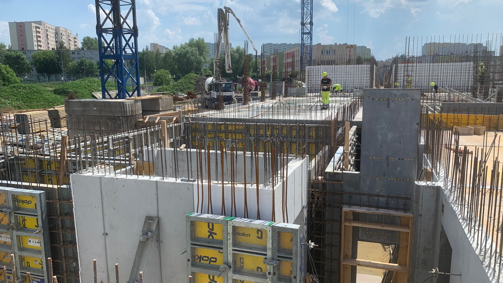 Wysoki kąt na budowę z betonowymi ścianami szalunkowymi, zbrojeniami i pracownikami w odblaskowych ubraniach. W tle widoczny dźwig i zabudowa miejska.