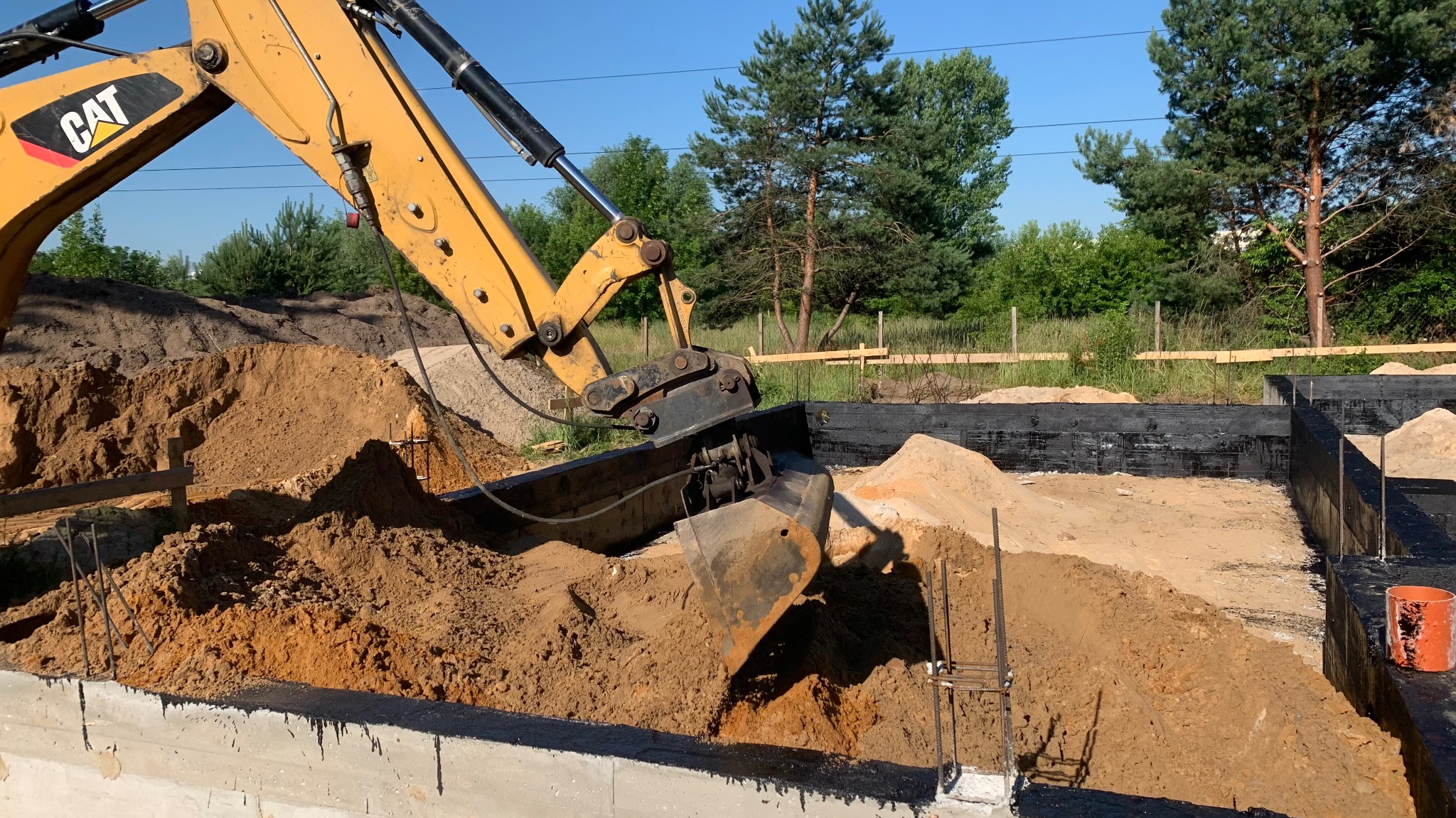 Żółta koparka CAT zasypuje fundament piaskiem. Widoczne zbrojenia i izolacja fundamentu. Prace ziemne w toku, błękitne niebo w tle.