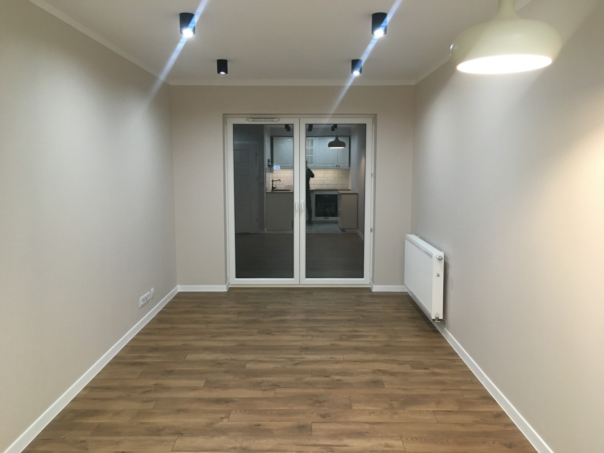 Jasne, puste pomieszczenie z panelami podłogowymi w odcieniach brązu, świeżo pomalowanymi ścianami w kolorze beżowym i widokiem na kuchnię przez przeszklone drzwi przesuwne.