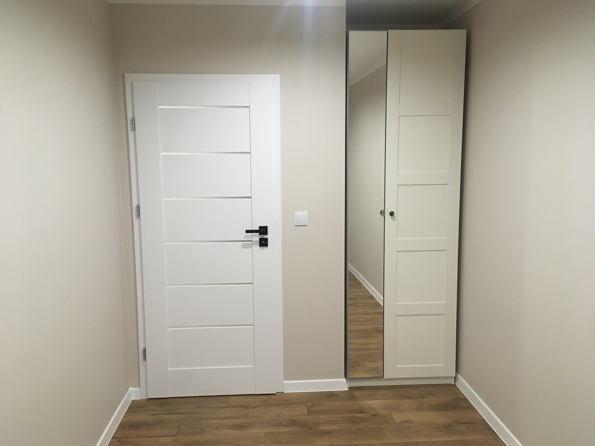 Białe, nowoczesne drzwi z czarną klamką obok szafy z lustrem i drewnianą podłogą w odcieniach brązu, świeżo po remoncie.