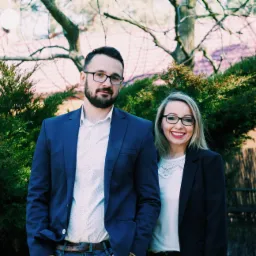 Uśmiechnięta para w eleganckich strojach na tle zieleni i budynku z czerwonej cegły, prawdopodobnie agenci nieruchomości.