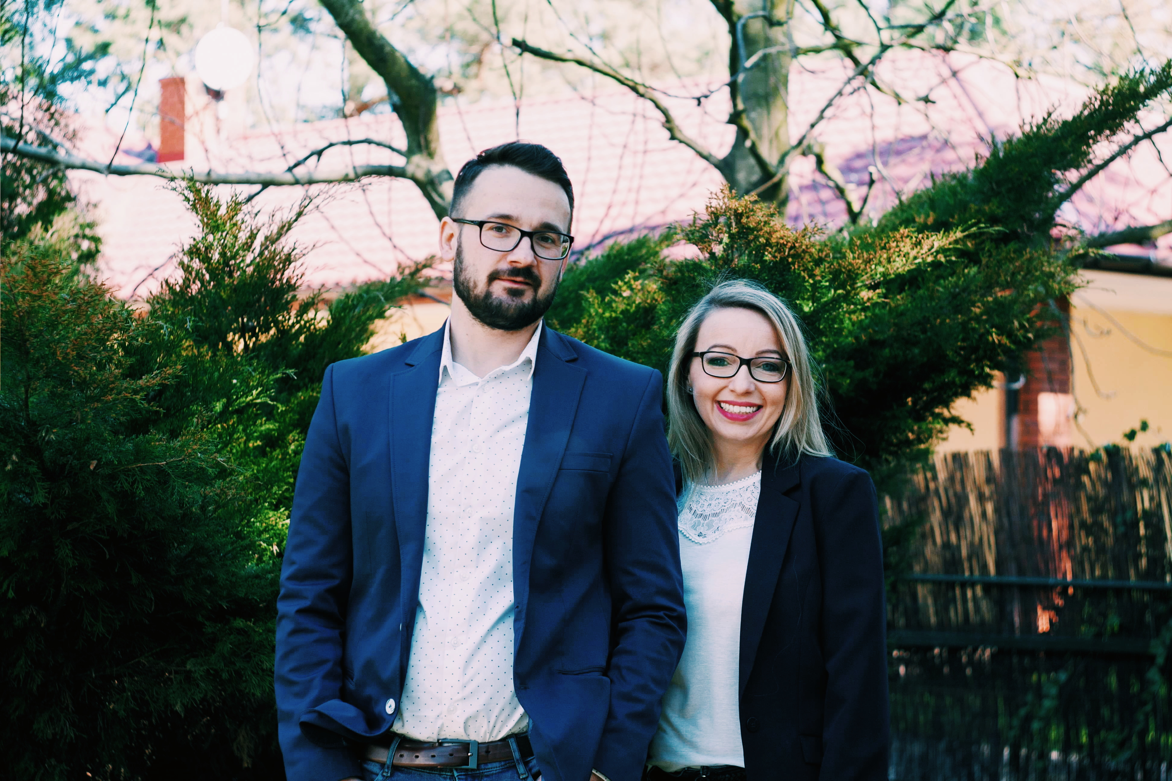 Uśmiechnięta para w eleganckich strojach na tle zieleni i budynku z czerwonej cegły, prawdopodobnie agenci nieruchomości.