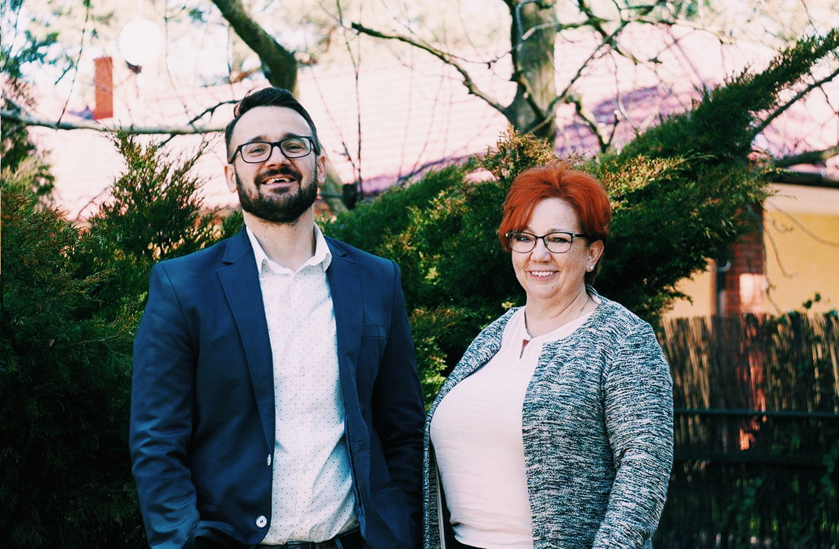 Portret uśmiechniętego mężczyzny w niebieskiej marynarce i kobiety w szarej narzutce, stojących przed zielonymi krzewami i drzewami.