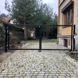 Dwuskrzydłowa brama wjazdowa z paneli stalowych, widoczna na tle budynku z oknami i brukowanej nawierzchni z mchem.