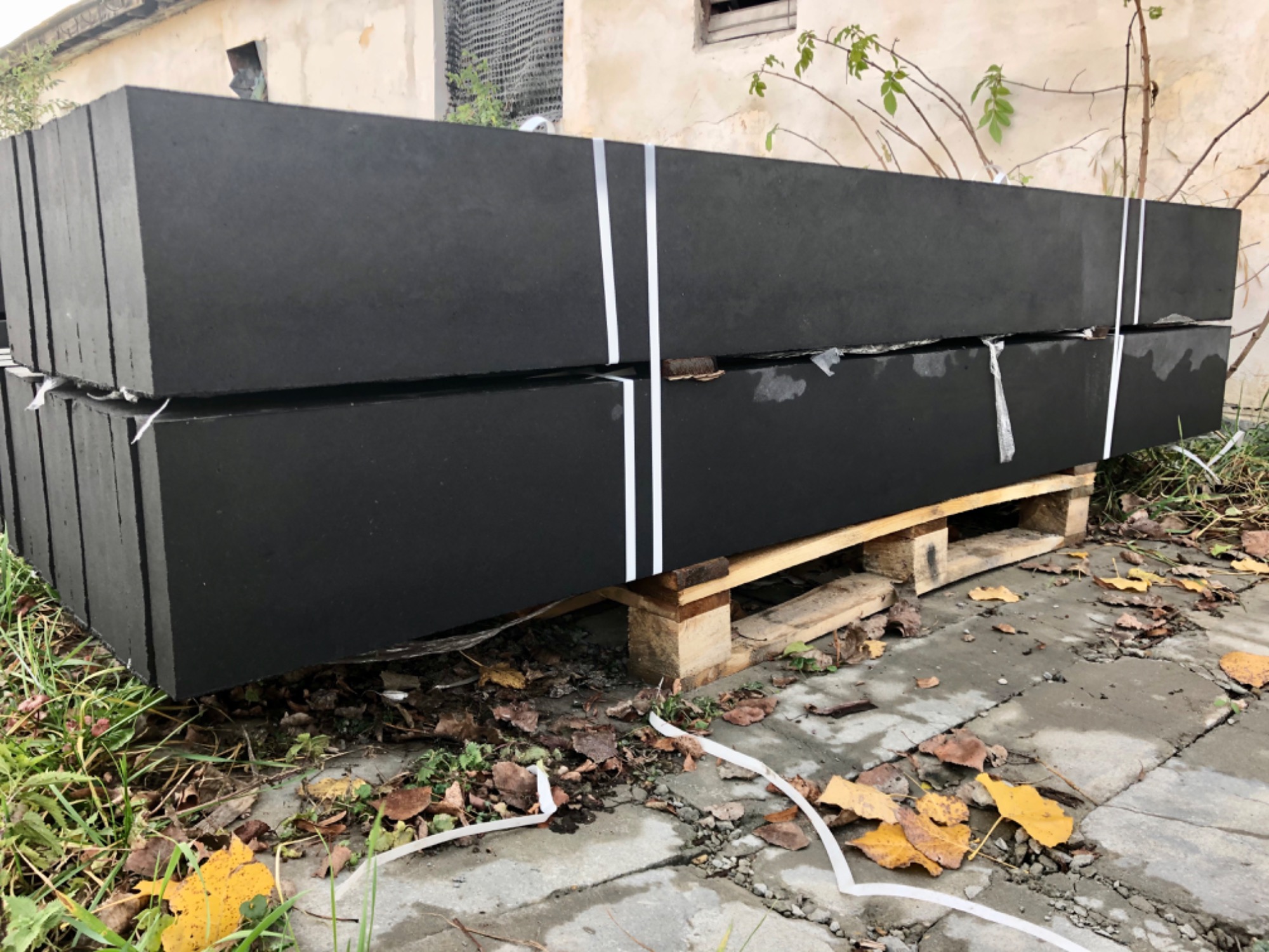Czarne, prefabrykowane elementy betonowe na palecie, owinięte białą taśmą, przygotowane do montażu ogrodzenia. Widoczne jesienne liście na ziemi.