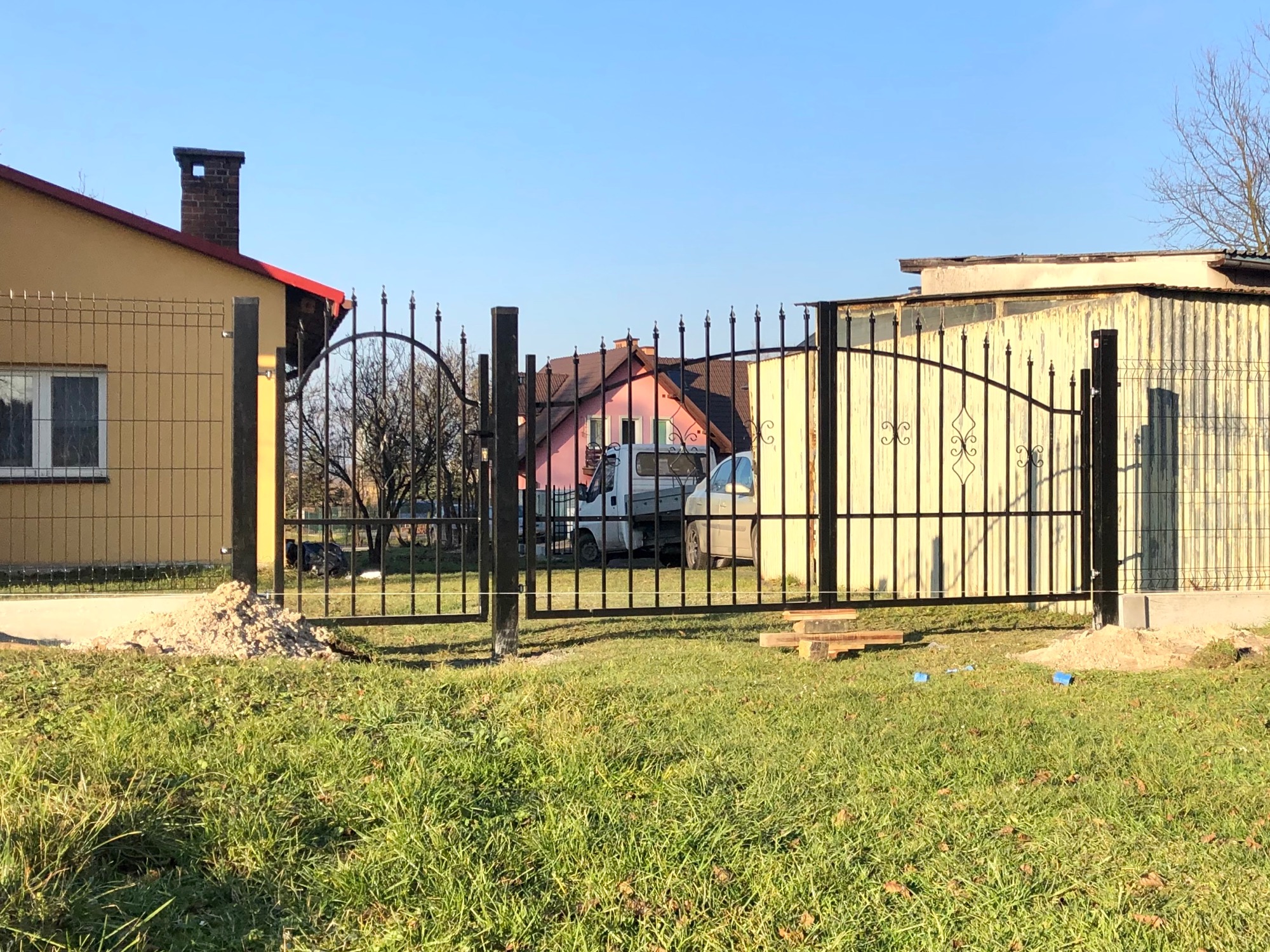 Częściowo zamontowana, czarna brama wjazdowa z ozdobnymi grotami, po lewej ogrodzenie panelowe, widoczny kopiec ziemi i elementy montażowe na trawie, w tle budynki mieszkalne i dostawczak.