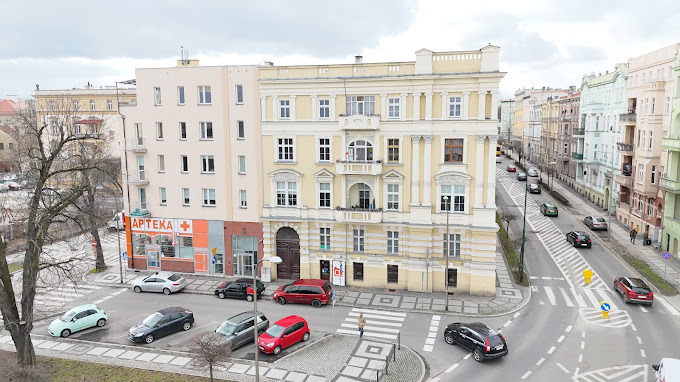 Widok z góry na skrzyżowanie ulic w mieście z budynkami o zróżnicowanej architekturze, w tym kamienica z balkonem i apteka na rogu, kilka samochodów na ulicy i pieszy na przejściu.