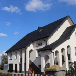 Dwupiętrowy dom z czarnym dachem z dachówki modułowej, jasną elewacją i kolumnami, widok z poziomu gruntu w słoneczny dzień.