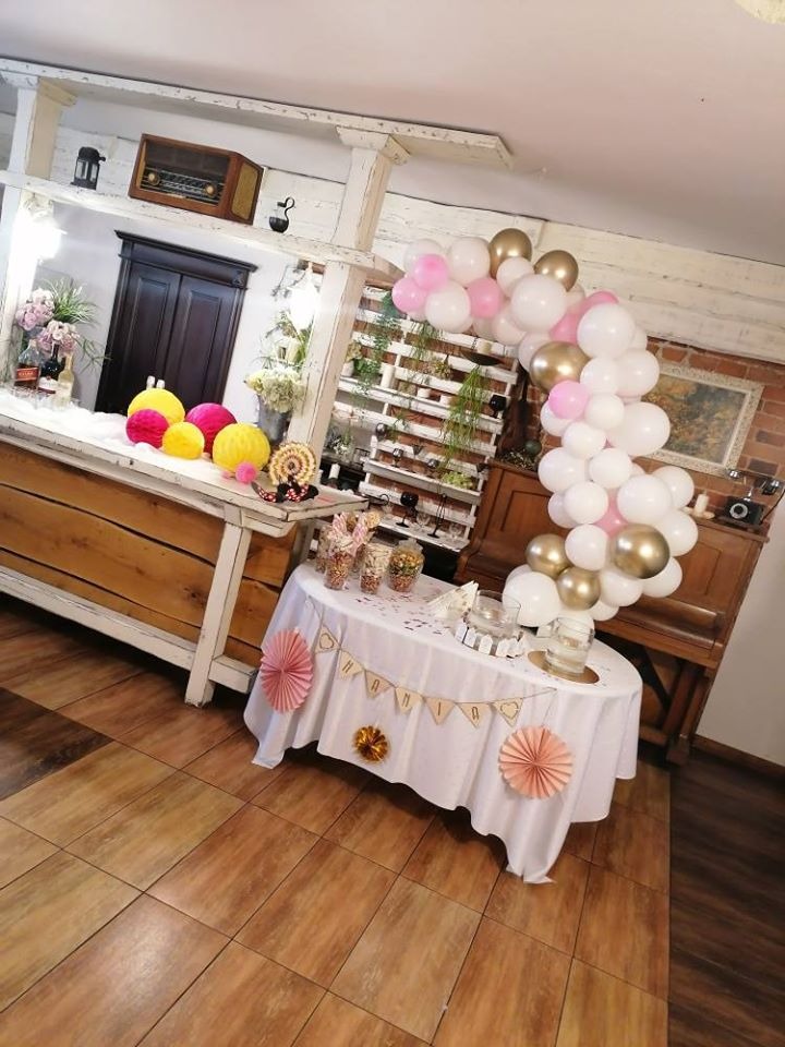 Stół udekorowany białym obrusem z dekoracjami papierowymi i girlandą, za nim łuk z białych, różowych i złotych balonów w rustykalnym wnętrzu.