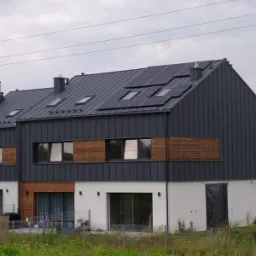 Nowoczesny szeregowiec z ciemną blachą na elewacji i panelami słonecznymi na dachu, widok z poziomu trawy w pochmurny dzień.