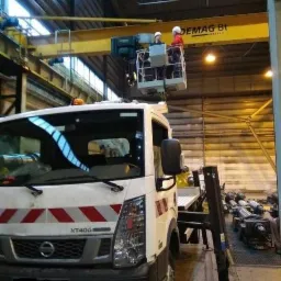 Podnośnik koszowy zamontowany na pojeździe Nissan NT400, z dwoma pracownikami w kasku, podwieszony pod suwnicą Demag 8t w hali przemysłowej.