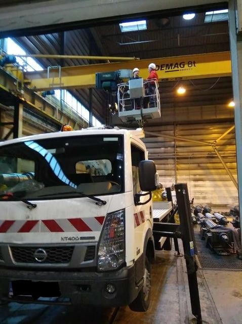 Podnośnik koszowy zamontowany na pojeździe Nissan NT400, z dwoma pracownikami w kasku, podwieszony pod suwnicą Demag 8t w hali przemysłowej.