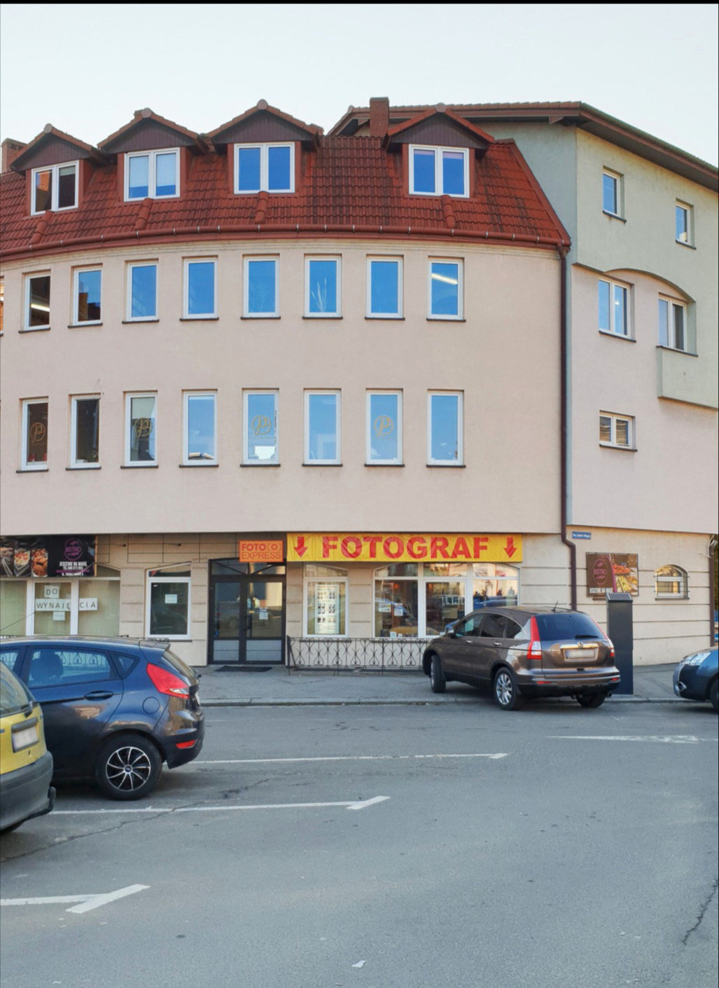 Fasada budynku z szyldem 'FOTOGRAF' nad wejściem, zaparkowane samochody na pierwszym planie.