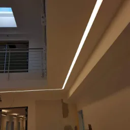 Wnętrze w trakcie wykańczania z zamontowanym liniowym oświetleniem LED w suficie podwieszanym, widoczna drabina malarska i fragment balkonu z metalową barierką.