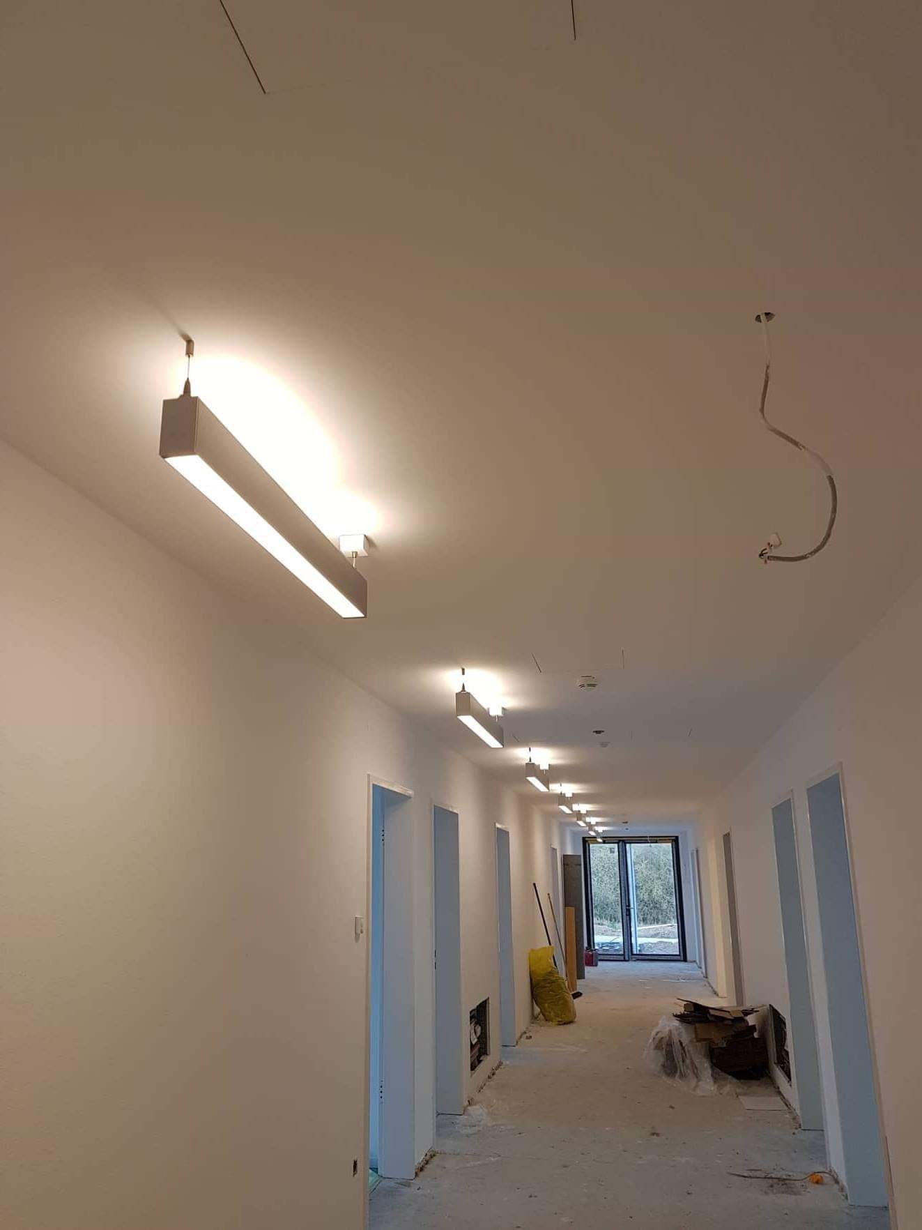 Długi korytarz w nowym budynku z zamontowanym liniowym oświetleniem sufitowym; widoczny przewód elektryczny zwisający z sufitu, sugerujący niedokończoną instalację.