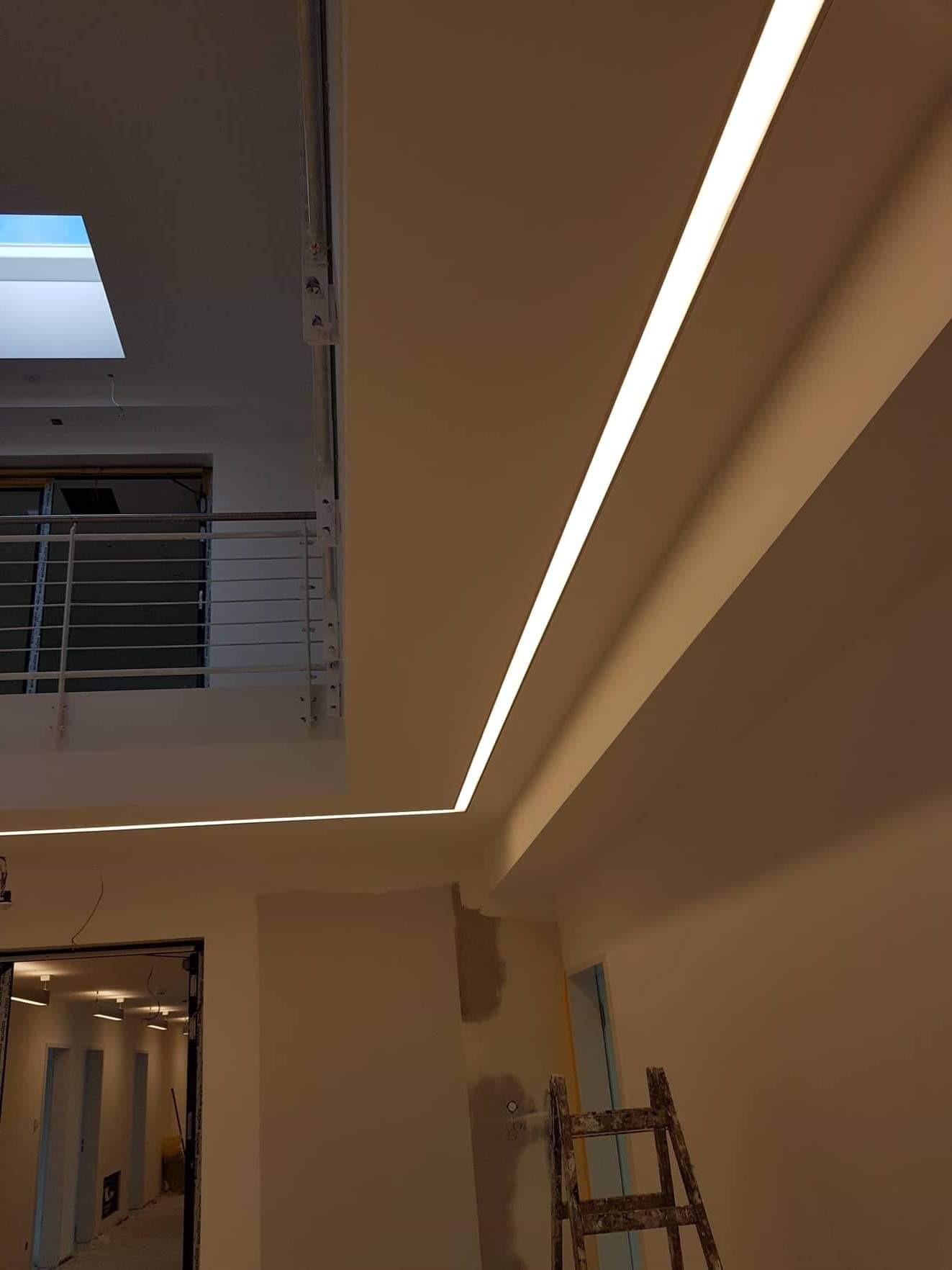 Wnętrze w trakcie wykańczania z zamontowanym liniowym oświetleniem LED w suficie podwieszanym, widoczna drabina malarska i fragment balkonu z metalową barierką.