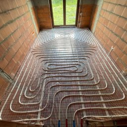 ENKAM SPÓŁKA Z OGRANICZONĄ ODPOWIEDZIALNOŚCIĄ - Instalacja ogrzewania podłogowego w surowym wnętrzu z ceglanymi ścianami. Białe rurki ułożone spiralnie na folii z siatką. Widok z góry na okno.