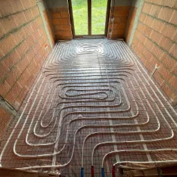 Instalacja ogrzewania podłogowego w surowym wnętrzu z ceglanymi ścianami. Białe rurki ułożone spiralnie na folii z siatką. Widok z góry na okno.