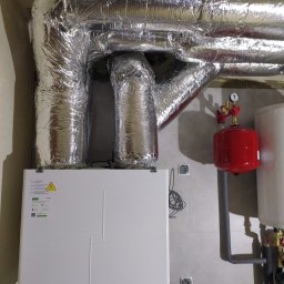 ENKAM SPÓŁKA Z OGRANICZONĄ ODPOWIEDZIALNOŚCIĄ - Biała centrala wentylacyjna Vents z izolowanymi, srebrnymi przewodami wentylacyjnymi, czerwonym zbiornikiem i osprzętem hydraulicznym na tle szarej ściany.